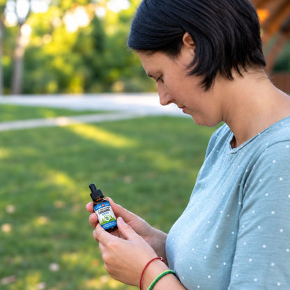 Donna in un parco che legge l'etichetta di un flacone di olio di CBD, maglietta azzurra a pois e braccialetti rossi e verdi